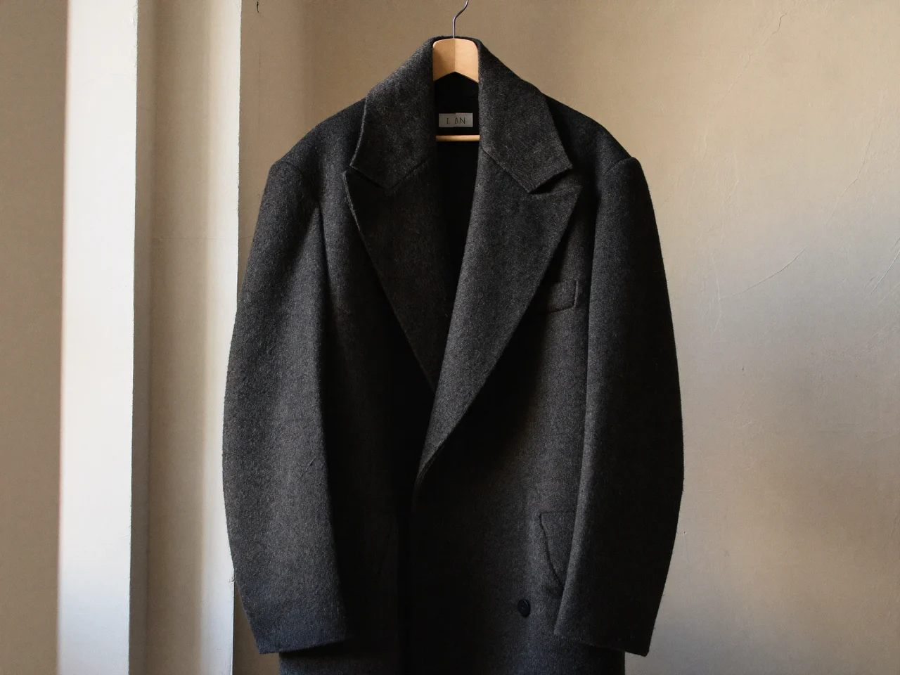 Cappotto oversize antracite appeso a una gruccia in legno davanti a un muro intonacato